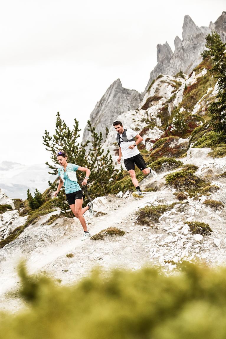 Deux coureurs de trail en plein effort sur un sentier rocheux &agrave; M&eacute;ribel, cr&ecirc;tes ac&eacute;r&eacute;es en arri&egrave;re-plan